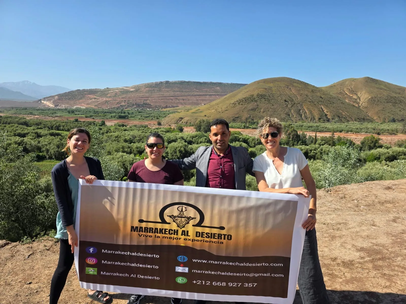 Guía de Marrakech Al Desierto con tres viajeras sujetando el cartel de la agencia en un valle del Alto Atlas, Marruecos